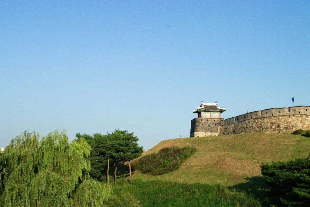 A stone corner tower and walls of Suwon Hwaseong Fortress on a green hillside under a clear blue sky